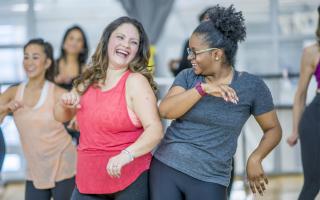 A group of young multi-ethnic women are participating in a Zumba class.  They are all wearing their active wear, smiling and having fun.  The two women in front are bumping hips and sharing a laugh.